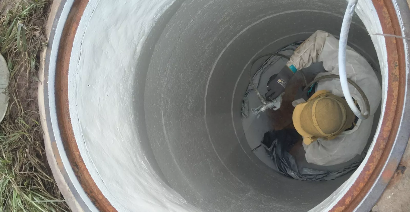 Technician in protective suit spraying seamless polyurea lining inside a municipal manhole using safety tripod setup
